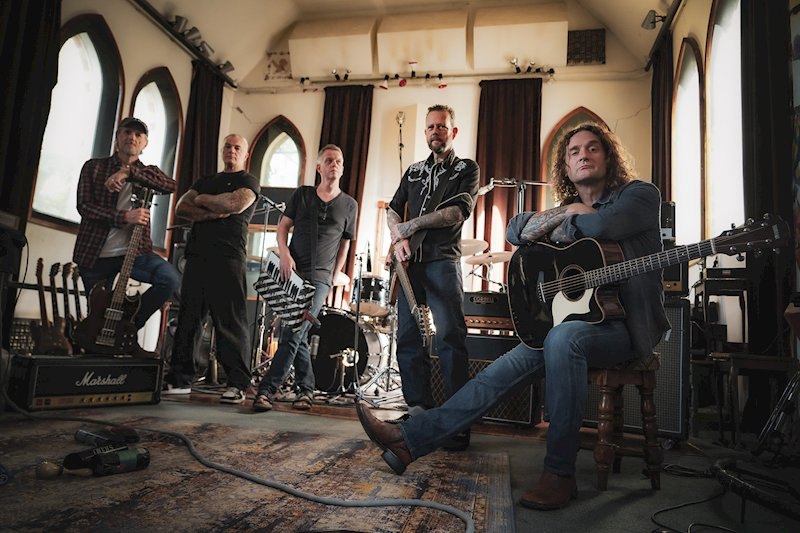 The five members of Little Angels in a studio with arched church windows, surrounded by amplifiers and guitars