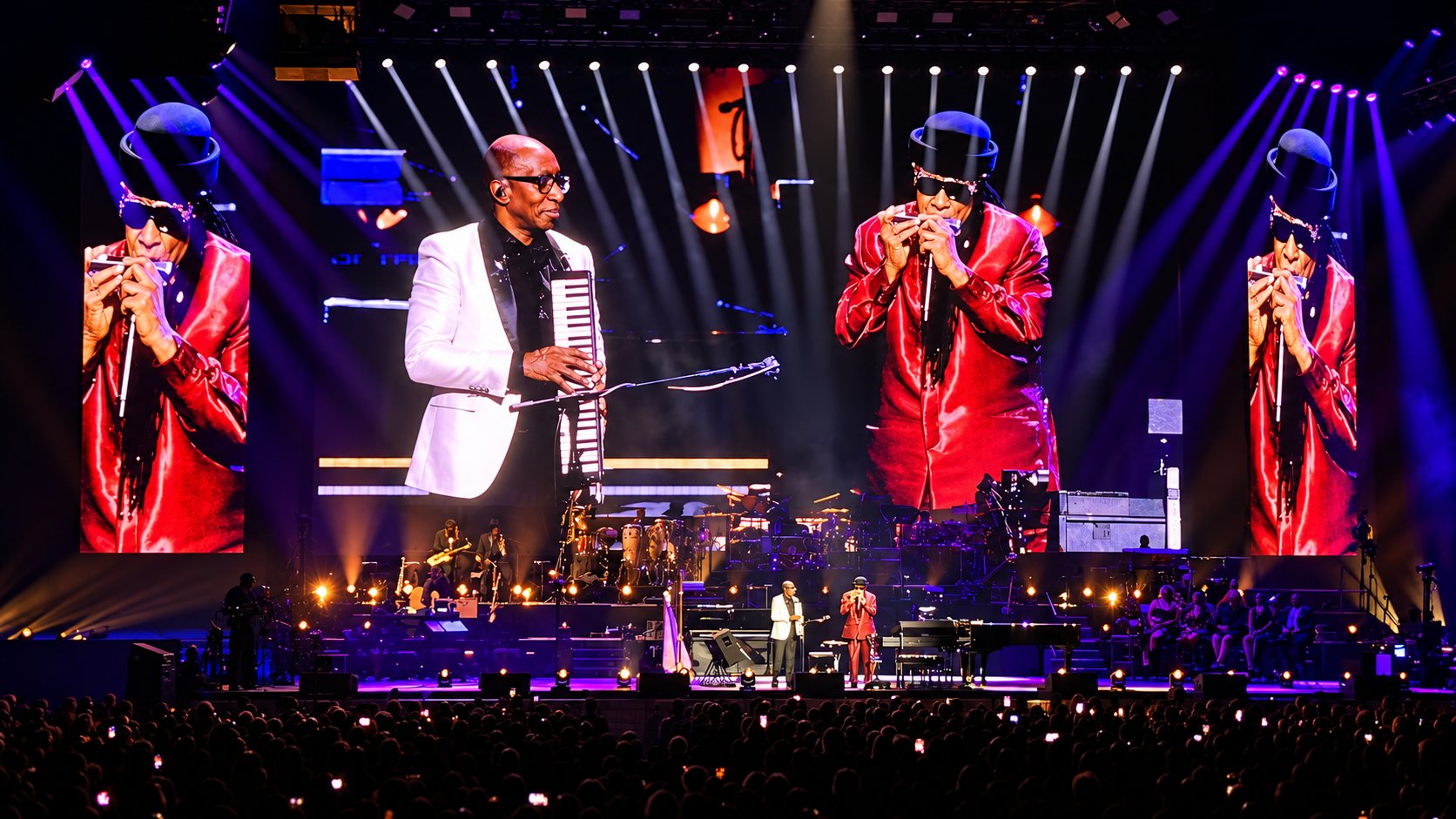 Stevie Wonder in a red jacket playing harmonica, shown on the giant screens above the stage of the Lanxess Arena, with a keyboard player in a white jacket beside him